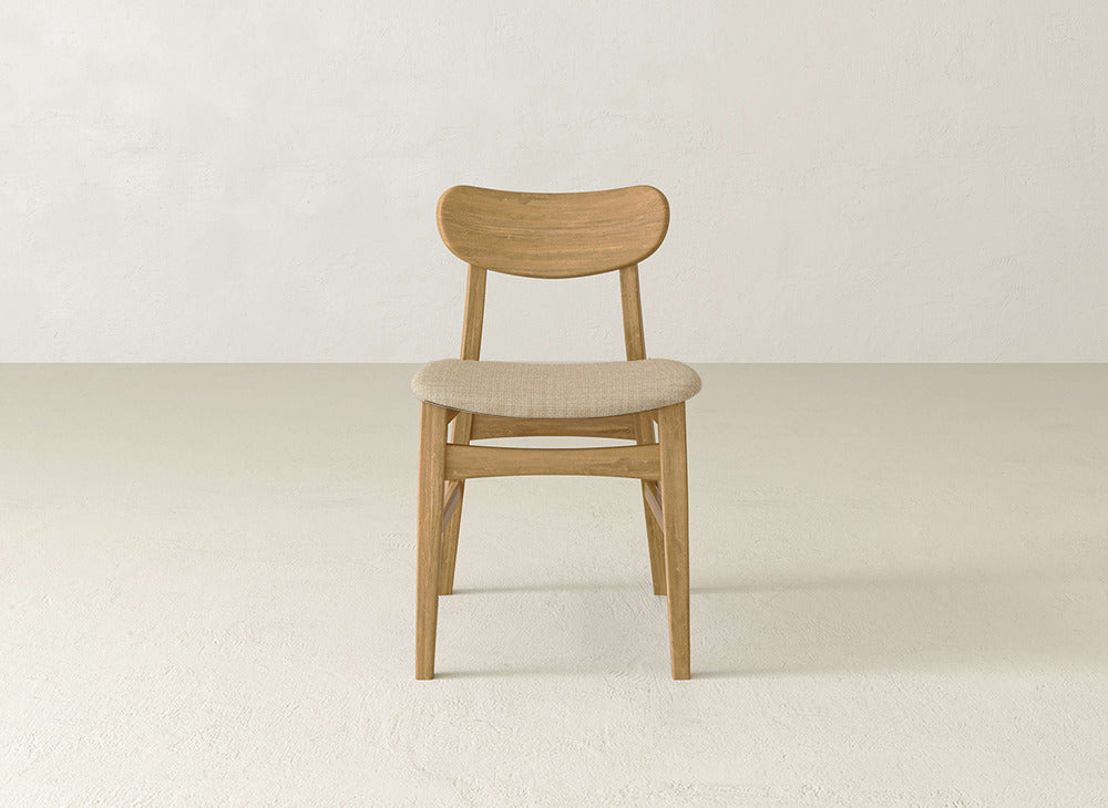 The Lynn Upholstered Dining Chair by James+James, featuring a curved maple wood backrest and a cushioned beige seat, sits on a light floor against a plain off-white wall.
