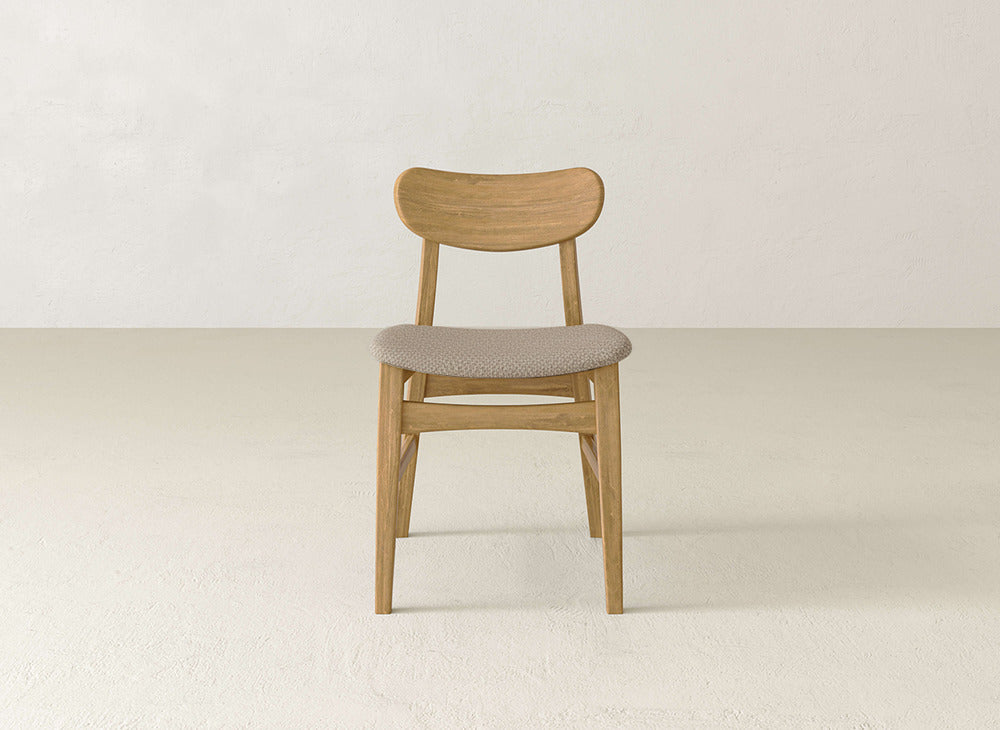 The Lynn Upholstered Dining Chair by James+James features a curved backrest and cushioned light beige seat, crafted from maple, and is showcased in a minimalist room with off-white walls and floors.