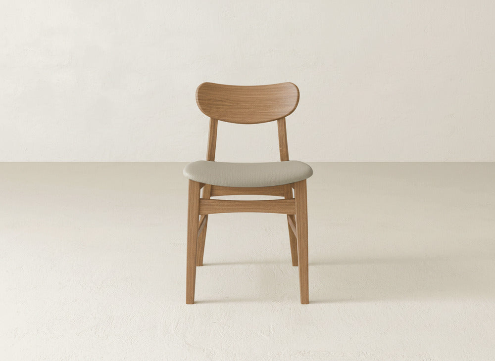 The Lynn Upholstered Dining Chair by James+James features a curved backrest and light gray cushioned seat, crafted from maple wood, adding modern sophistication to any minimal, light-colored room with a smooth floor and plain wall.