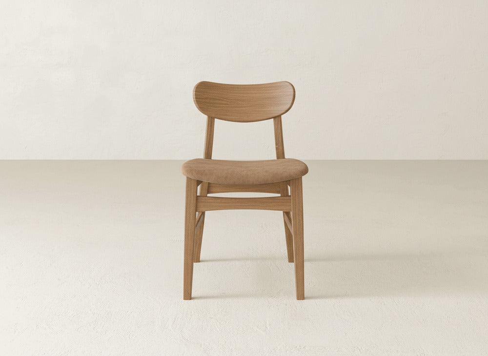 A Lynn Upholstered Dining Chair by James+James, featuring a curved backrest and rounded seat in a minimalist, mid-century modern maple wood design, sits on a light textured floor against an off-white wall.