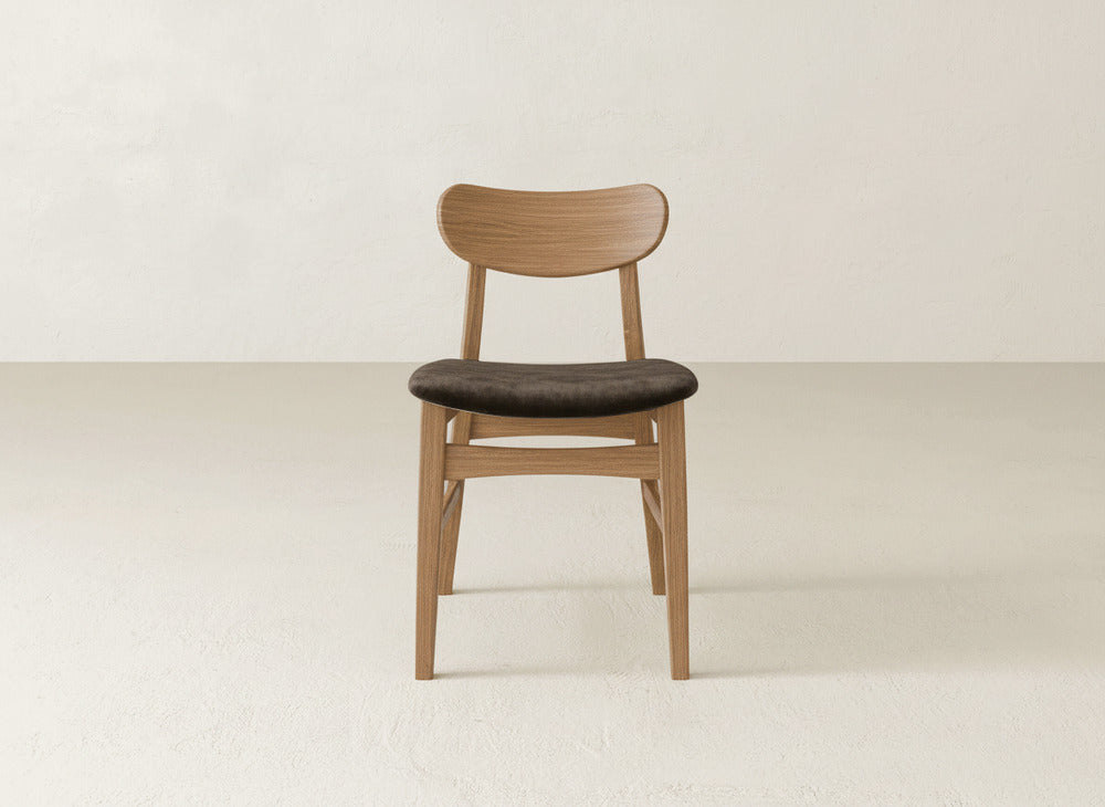 The Lynn Upholstered Dining Chair by James+James showcases a curved maple wood backrest and dark cushioned seat, placed on a light floor with an off-white wall backdrop.