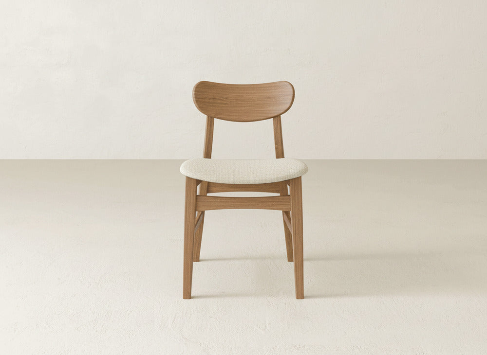 The Lynn Upholstered Dining Chair by James+James features a curved maple wood backrest and light cushioned seat, adding modern elegance to any space. Shown here on a textured floor against an off-white wall.