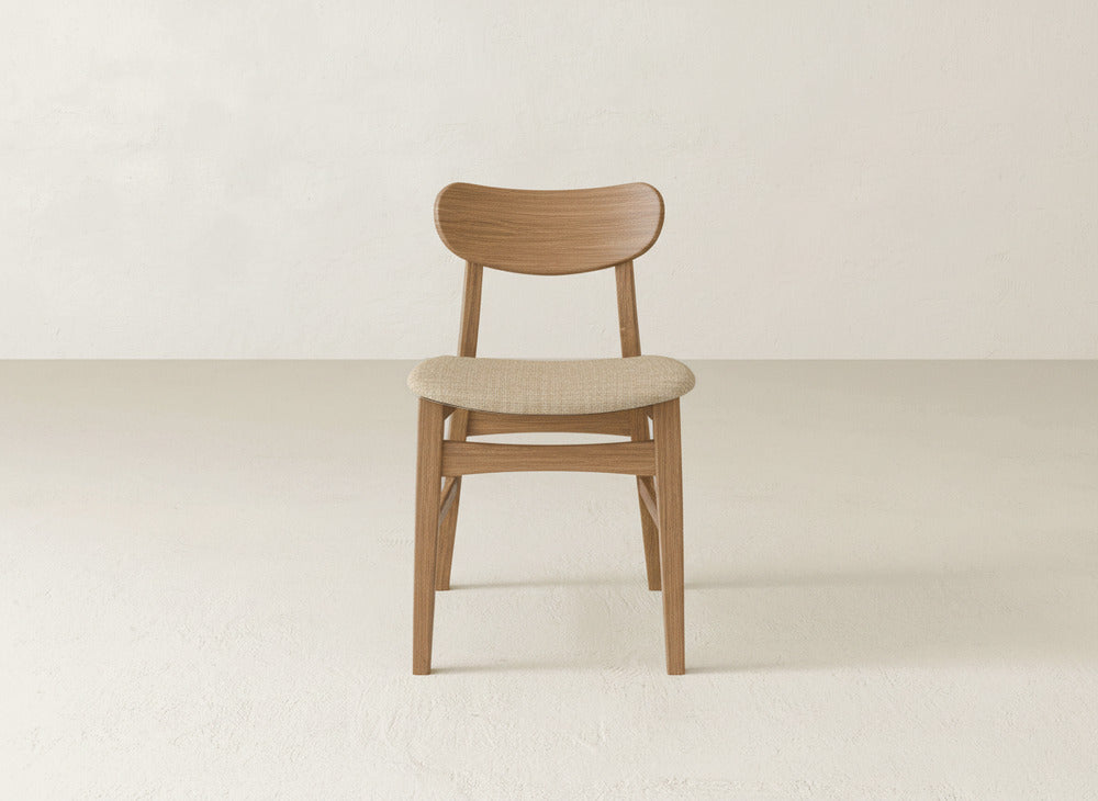 The Lynn Upholstered Dining Chair by James+James showcases modern style with a curved maple wood backrest and beige seat, displayed on a light floor against an off-white wall.