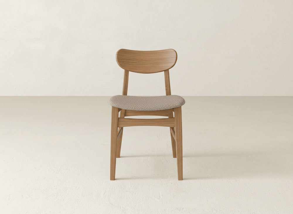 The Lynn Upholstered Dining Chair by James+James features a curved backrest and light beige seat, crafted from maple wood and set in the center of a minimalist, light-toned room with plain walls and floor.