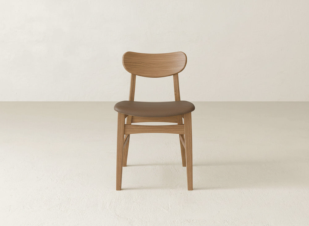 The Lynn Upholstered Dining Chair by James+James features a curved backrest and smooth, dark brown maple wood seat, radiating modern style when placed on a light floor against an off-white wall.