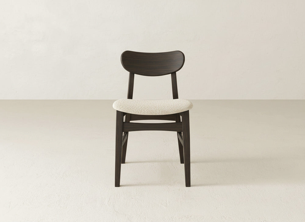 The Lynn Upholstered Dining Chair by James+James features a dark, curved maple wood backrest and legs, paired with a light, textured upholstered seat. It stands on a light floor against a plain off-white wall.