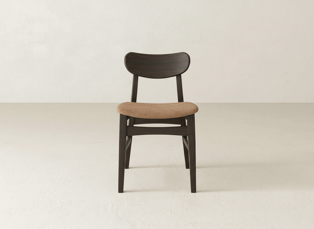 The Lynn Upholstered Dining Chair by James+James features a curved backrest and light brown cushioned seat, crafted from rich maple wood. This modern chair is shown on a light floor against an off-white wall.