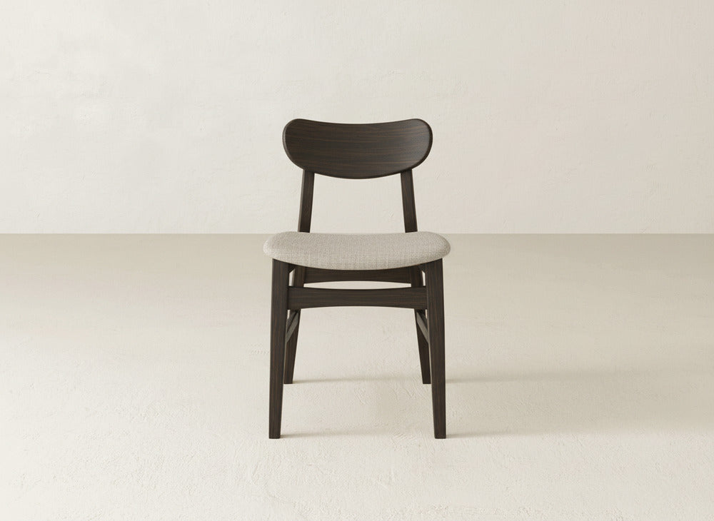The Lynn Upholstered Dining Chair by James+James features a dark maple wood frame, curved backrest, and light gray cushioned seat, shown on a plain light floor with an off-white wall in the background.