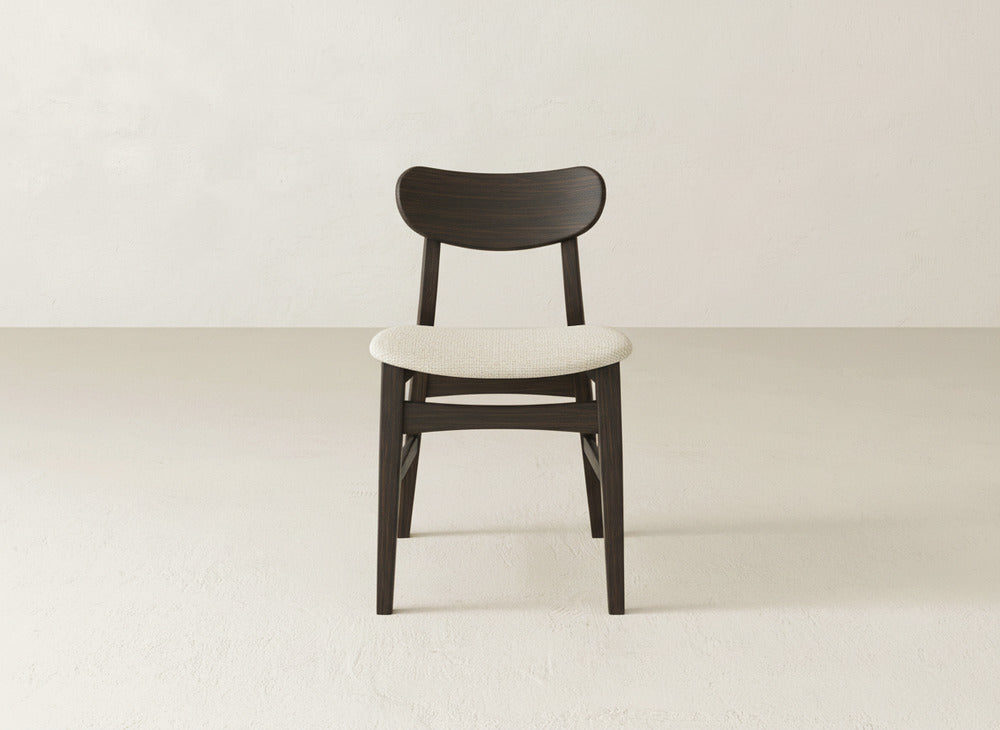 The Lynn Upholstered Dining Chair by James+James features a curved maple backrest, dark brown legs, and a light beige cushioned seat, bringing modern sophistication to rooms with light floors and walls.