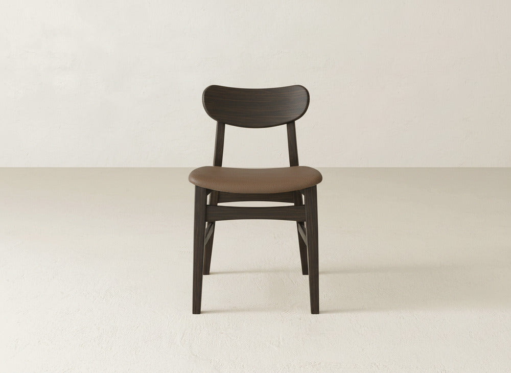 The Lynn Upholstered Dining Chair by James+James features a curved backrest and brown cushion, crafted from dark maple wood, adding modern elegance as it stands in the center of a bright room with a textured floor.