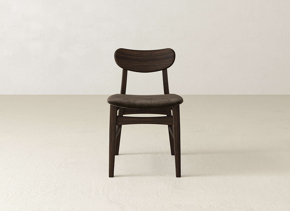 The Lynn Upholstered Dining Chair by James+James boasts a dark maple frame, curved backrest, and cushioned seat, blending modern elegance with comfort—perfect on a light floor beside an off-white wall.
