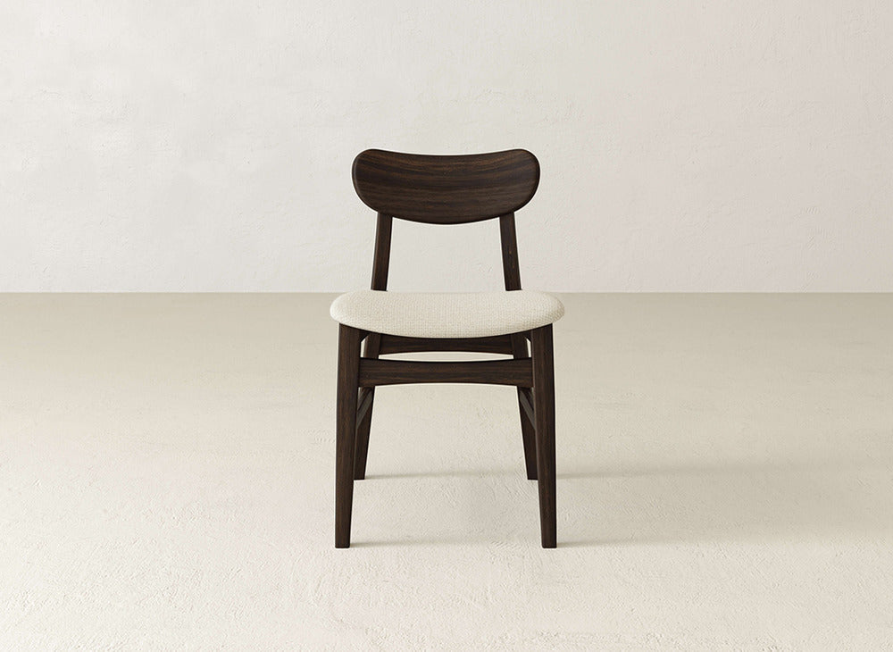 The Lynn Upholstered Dining Chair by James+James features a curved backrest and light cushioned seat made from maple wood, shown on a simple light floor with a neutral wall background.