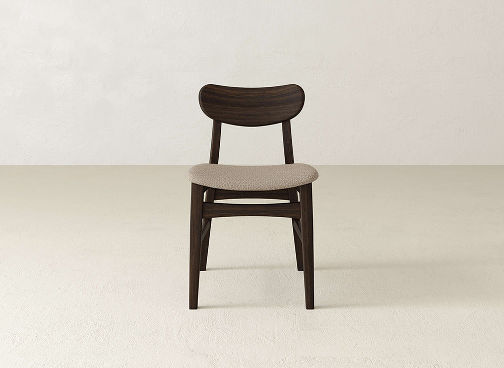 The Lynn Upholstered Dining Chair by James+James is a mid-century modern piece crafted from maple with a curved backrest and light beige upholstered seat, shown on a light floor against a neutral wall.