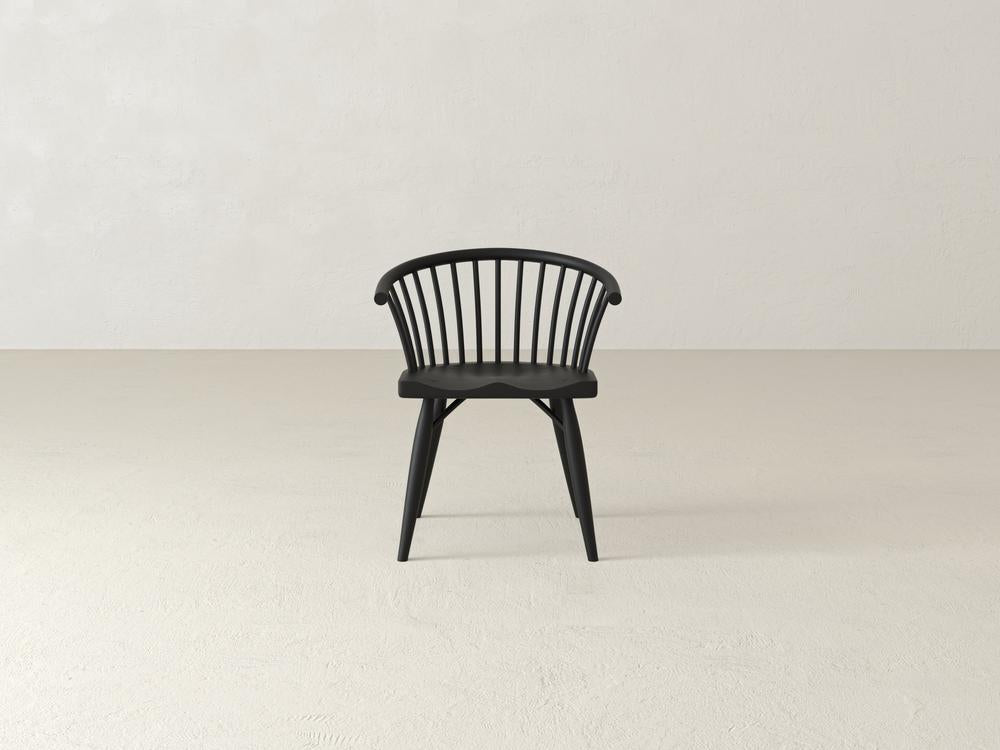 A wooden chair with a curved backrest and vertical slats stands on a plain, light-colored floor against a neutral wall background. The chair has tapered legs and an elegant, minimalist design.