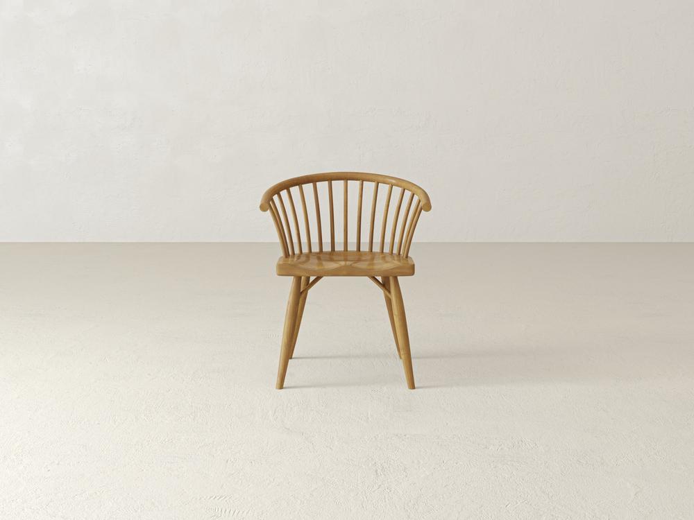 A wooden chair with a curved backrest and vertical slats sits on a plain, light-colored floor against a neutral wall. The design is minimalist, showcasing a smooth finish and tapered legs.