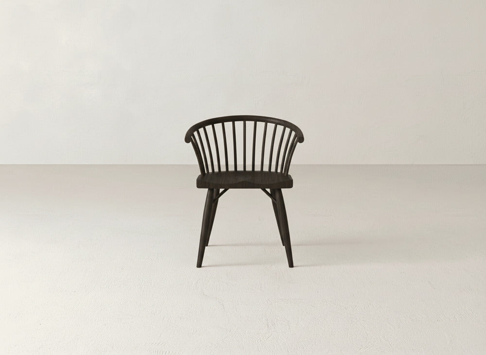 The Maria Dining Chair by James+James, featuring a dark wood finish, curved backrest, and vertical slats, stands alone in a minimalist room with light walls and a bare floor.