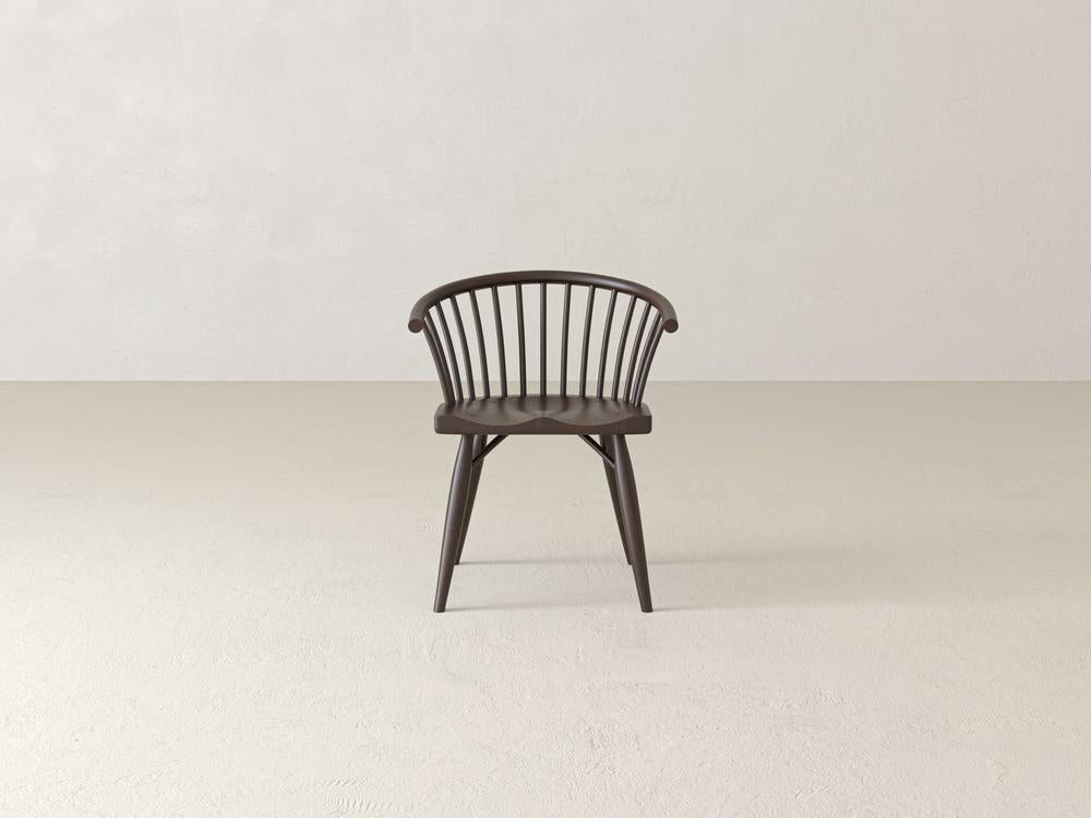 Dark wooden chair with spindle backrest, curved arms, and angled legs placed on a light, textured floor in a minimalist setting.