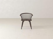Dark wooden chair with spindle backrest, curved arms, and angled legs placed on a light, textured floor in a minimalist setting.