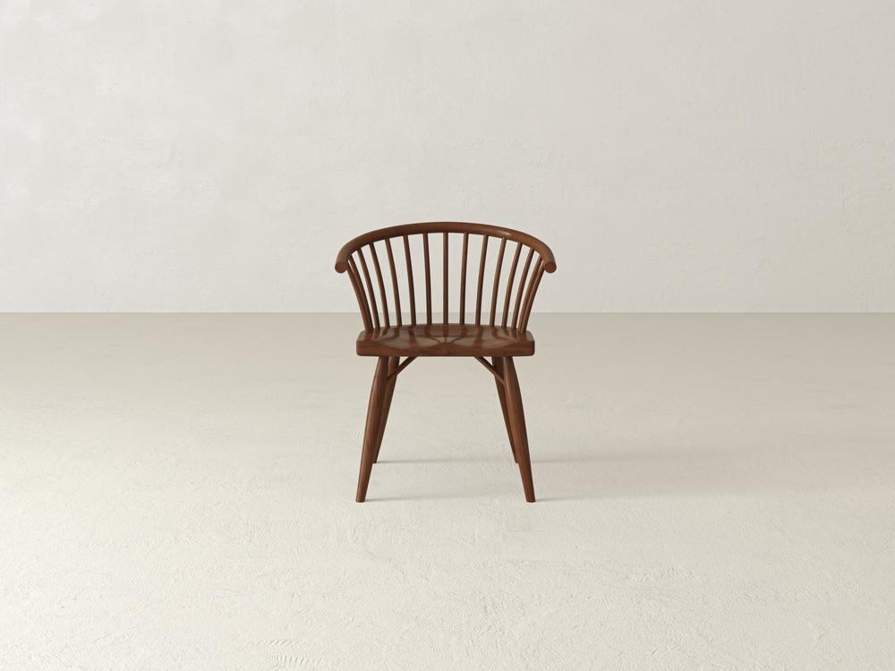 A wooden chair with a curved backrest and vertical slats sits on a plain, light-colored floor against a neutral wall. The design is minimalist, showcasing a smooth finish and tapered legs.
