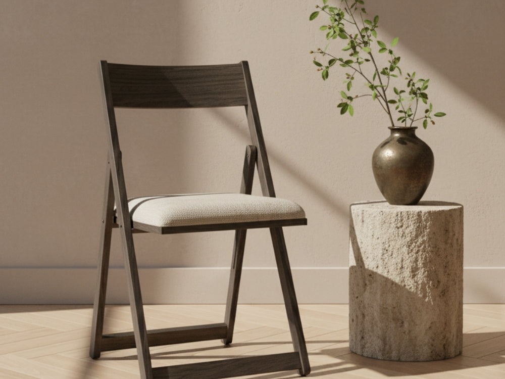 A James+James folding chair with a Natural - Linen Swatch seat sits by a round stone table with a vase of green branches. Sunlight casts shadows across the light wall and wooden floor.