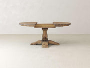 A wooden round table with an unfinished surface, displaying a central pedestal base and four evenly spaced legs. The background is a plain off-white wall and floor, creating a minimalist setting.