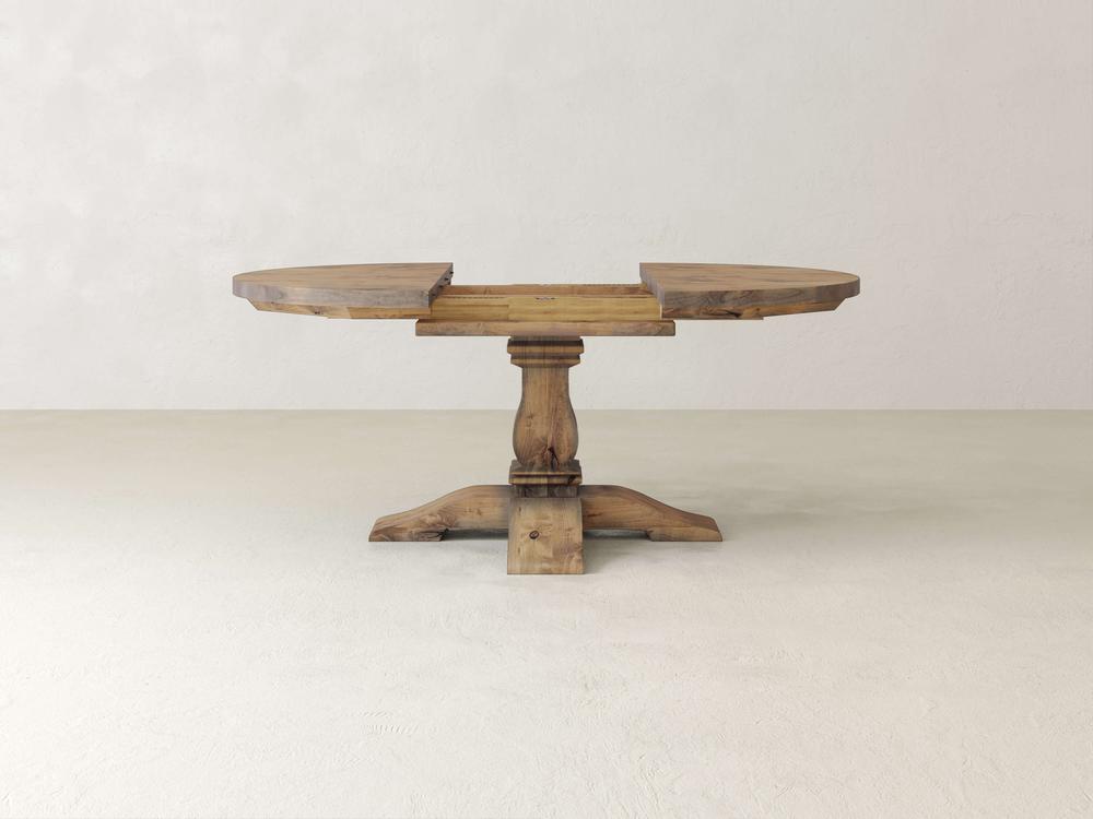 A wooden round table with an unfinished surface, displaying a central pedestal base and four evenly spaced legs. The background is a plain off-white wall and floor, creating a minimalist setting.