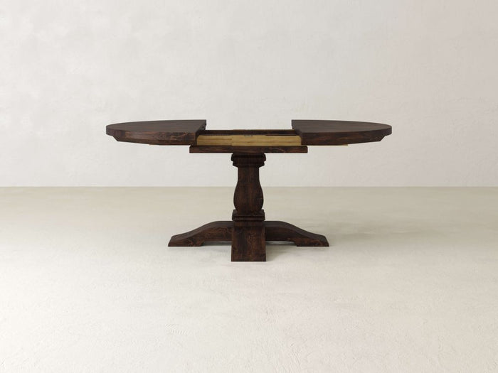 The Expandable Round Heirloom Dining Table by James & James, featuring a Knotty Alder tabletop and pedestal base, is partially extended with visible 2' L leaves and rests on a plain, light-colored floor against a white wall.