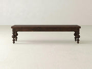 The Olivia Bench - Tobacco on Alder by James+James features a long, dark wooden rectangular top with four intricately carved, rounded legs, set on a light textured floor against a plain light wall.