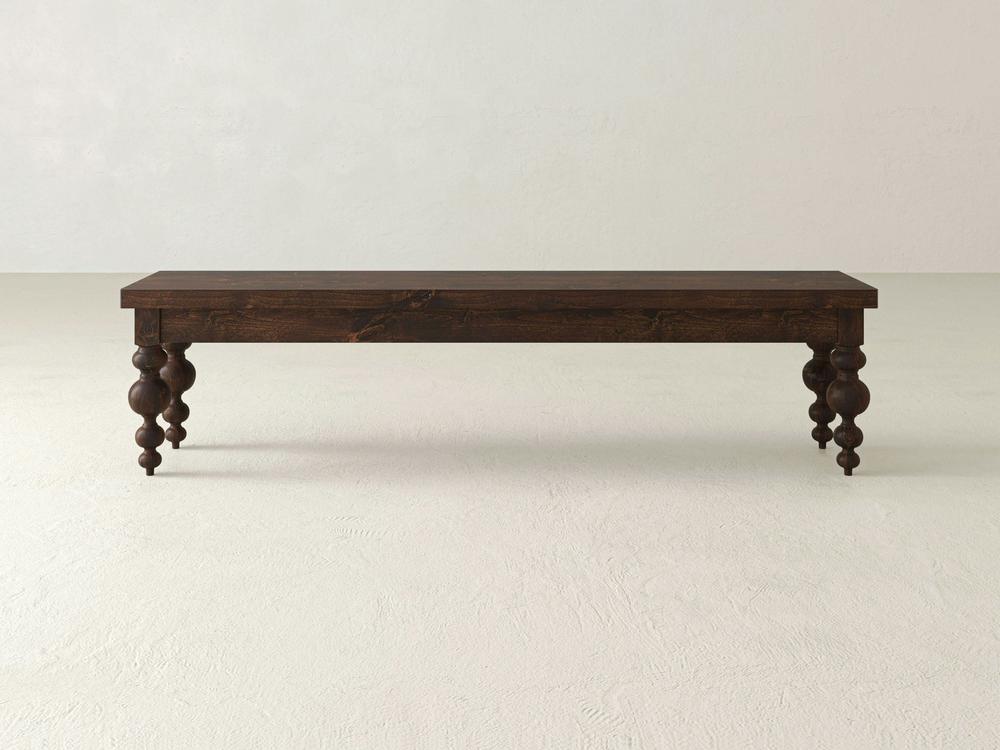 The Olivia Bench - Tobacco on Alder by James+James features a long, dark wooden rectangular top with four intricately carved, rounded legs, set on a light textured floor against a plain light wall.