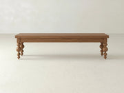The Olivia Bench - Almond on Oak by James+James features a rectangular wooden top and four rounded spindle-style legs, displayed against a plain, light-colored wall and floor.