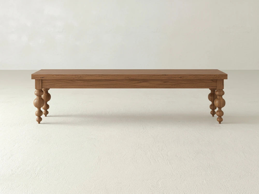 The Olivia Bench - Almond on Oak by James+James features a rectangular wooden top and four rounded spindle-style legs, displayed against a plain, light-colored wall and floor.