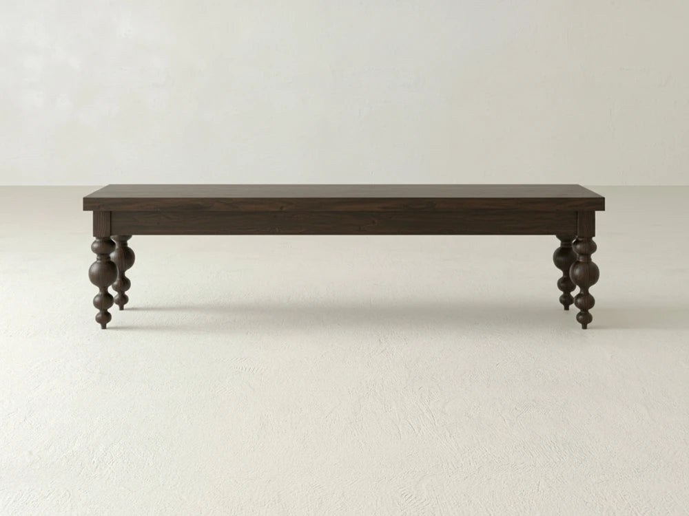 The Olivia Bench - Sable on Oak by James+James features dark wood and ornate, rounded legs, standing on a light floor against a plain off-white wall.