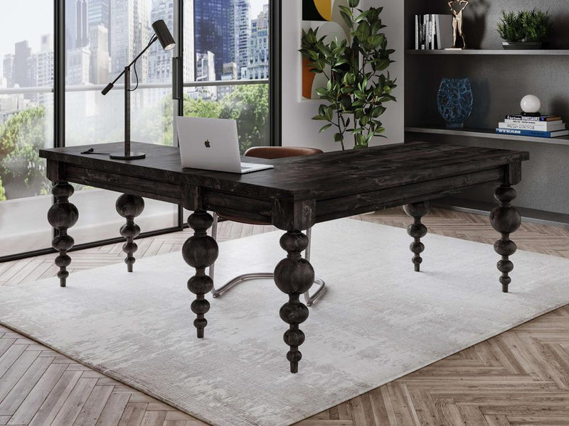 A modern office setup featuring the sturdy "Olivia Corner Desk - Charred Ember" by James+James with ornate legs. A laptop, lamp, and book sit on the desk. The floor has a light-colored rug and herringbone wood pattern. Large windows offer a city view, and a plant and shelves provide additional decor.