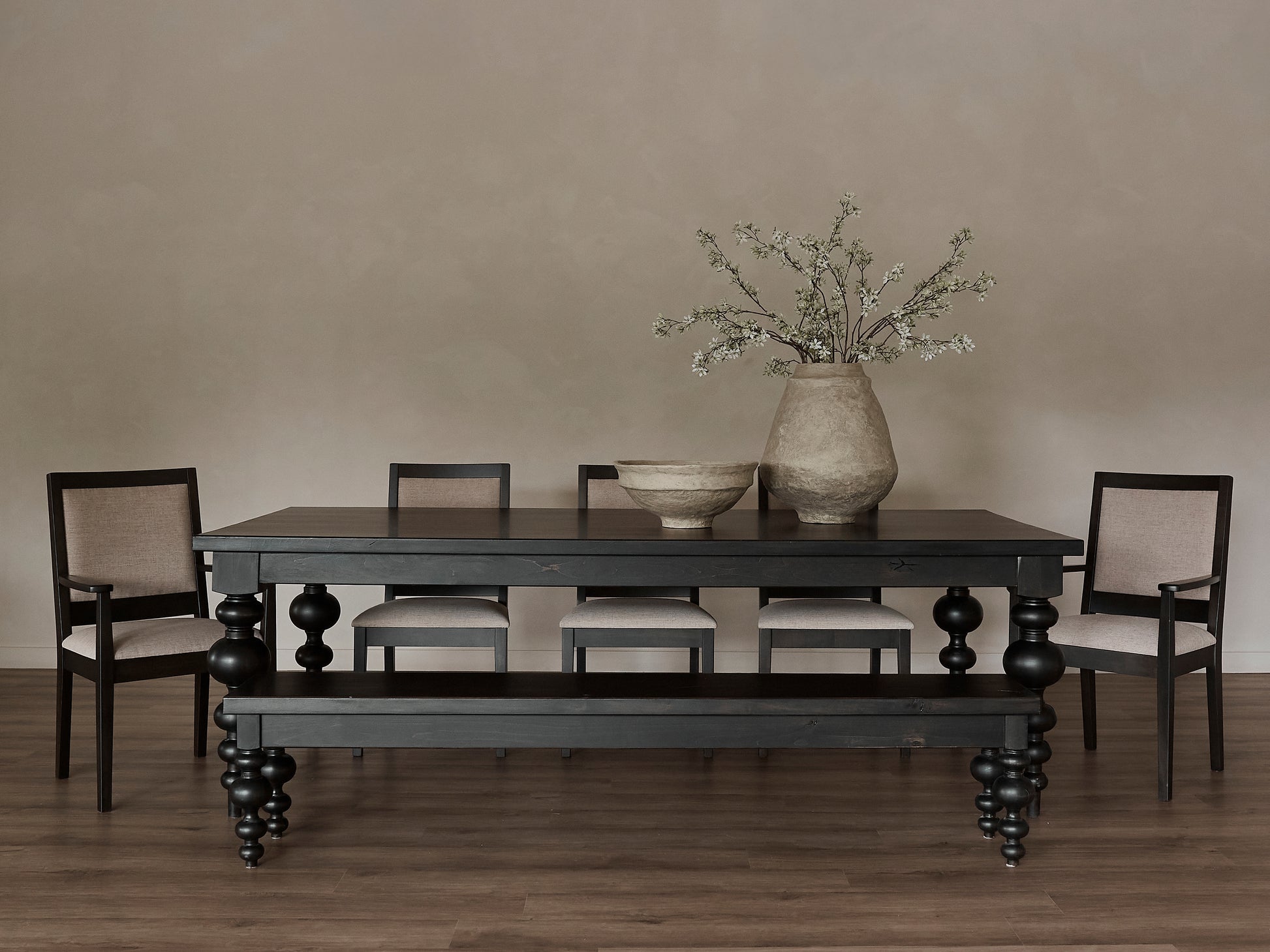 A minimalist dining room features the James+James Olivia Dining Table in Charred Ember with four beige-cushioned chairs and two large ceramic vases—one filled with white-blossomed branches—set against neutral walls and wooden floors.