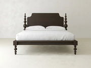 The Olivia Poster Bed by James+James features carved spindle posts, a solid headboard, and an espresso on oak finish, shown with a white mattress and two pillows against a light-colored wall and floor.