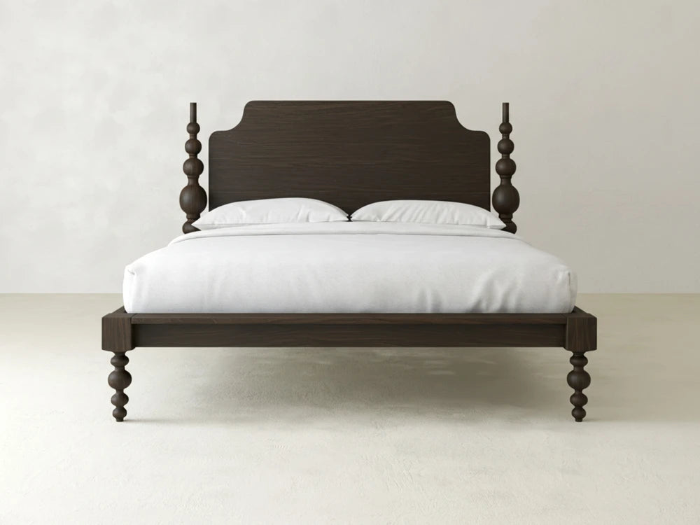 The Olivia Poster Bed by James+James features carved spindle posts, a solid headboard, and an espresso on oak finish, shown with a white mattress and two pillows against a light-colored wall and floor.