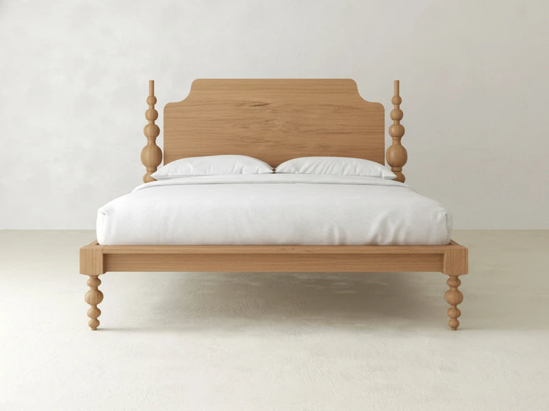 The Olivia Poster Bed - Oat on Oak by James+James features a wooden frame with a matching headboard, four turned legs, white bedding, and two pillows, set against a plain light-colored wall and floor.