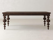 The Olivia Dining Table - Tobacco by James+James is a rectangular wooden table featuring intricately carved, spherical legs in a dark finish. It is set against a light-colored wall and rests on a smooth, light-toned floor.
