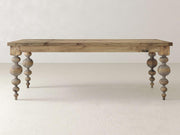 The Olivia Dining Table - Harvest Wheat by James+James features intricately turned legs and stands on a plain, light-colored floor. The tabletop boasts a natural finish, beautifully highlighting the wood grain. In the background is a smooth, neutral-colored wall.