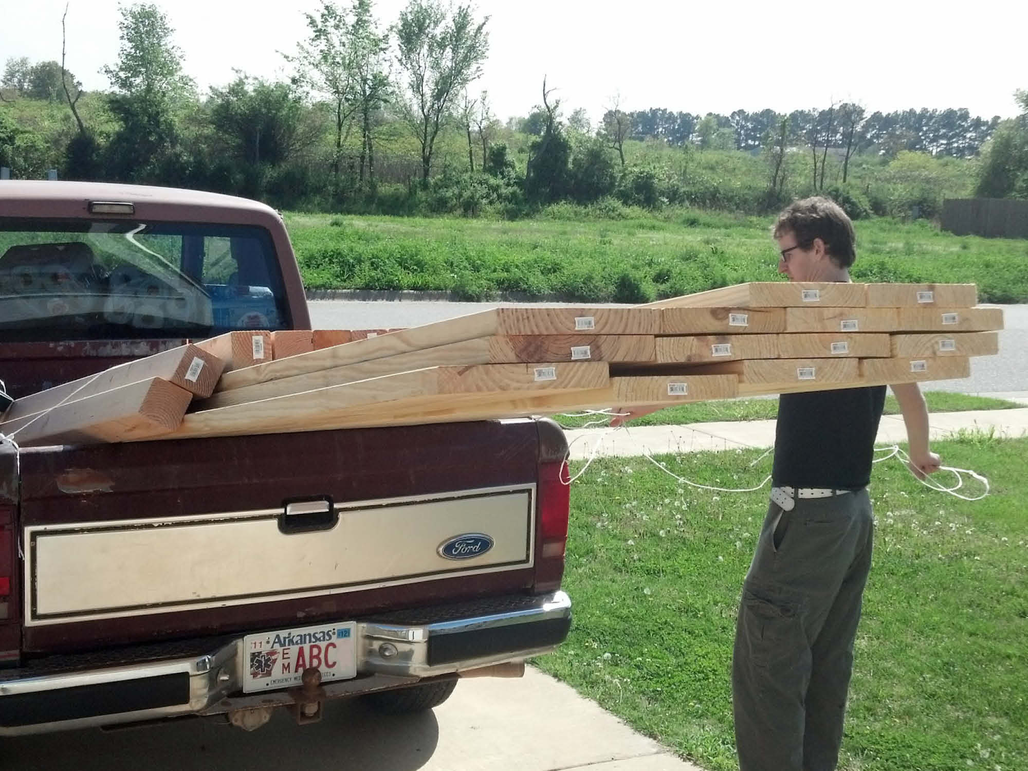 A person unloads long wooden planks from the back of a red Ford pickup truck parked on a driveway. The truck has an Arkansas license plate. The scene is set outdoors with grass and trees in the background.