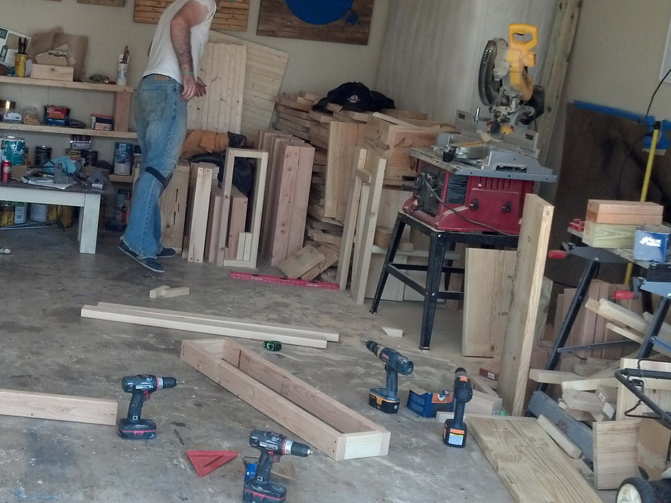 A person stands in a cluttered woodworking shop, surrounded by wooden boards, tools, and a circular saw. Several drills and a measuring square are on the floor. Shelves at the back hold more wood and supplies.