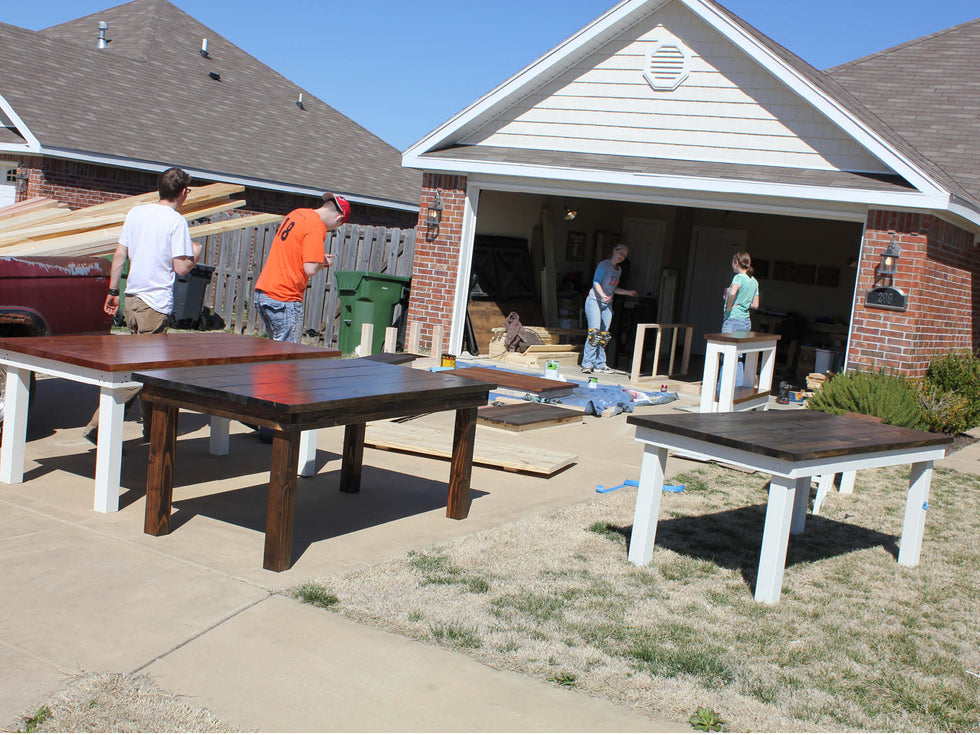 Four people are working on wooden tables outdoors near a brick garage. Three dark wooden tables with white legs are displayed on the driveway, and various tools and materials are scattered around. The sky is clear and sunny.