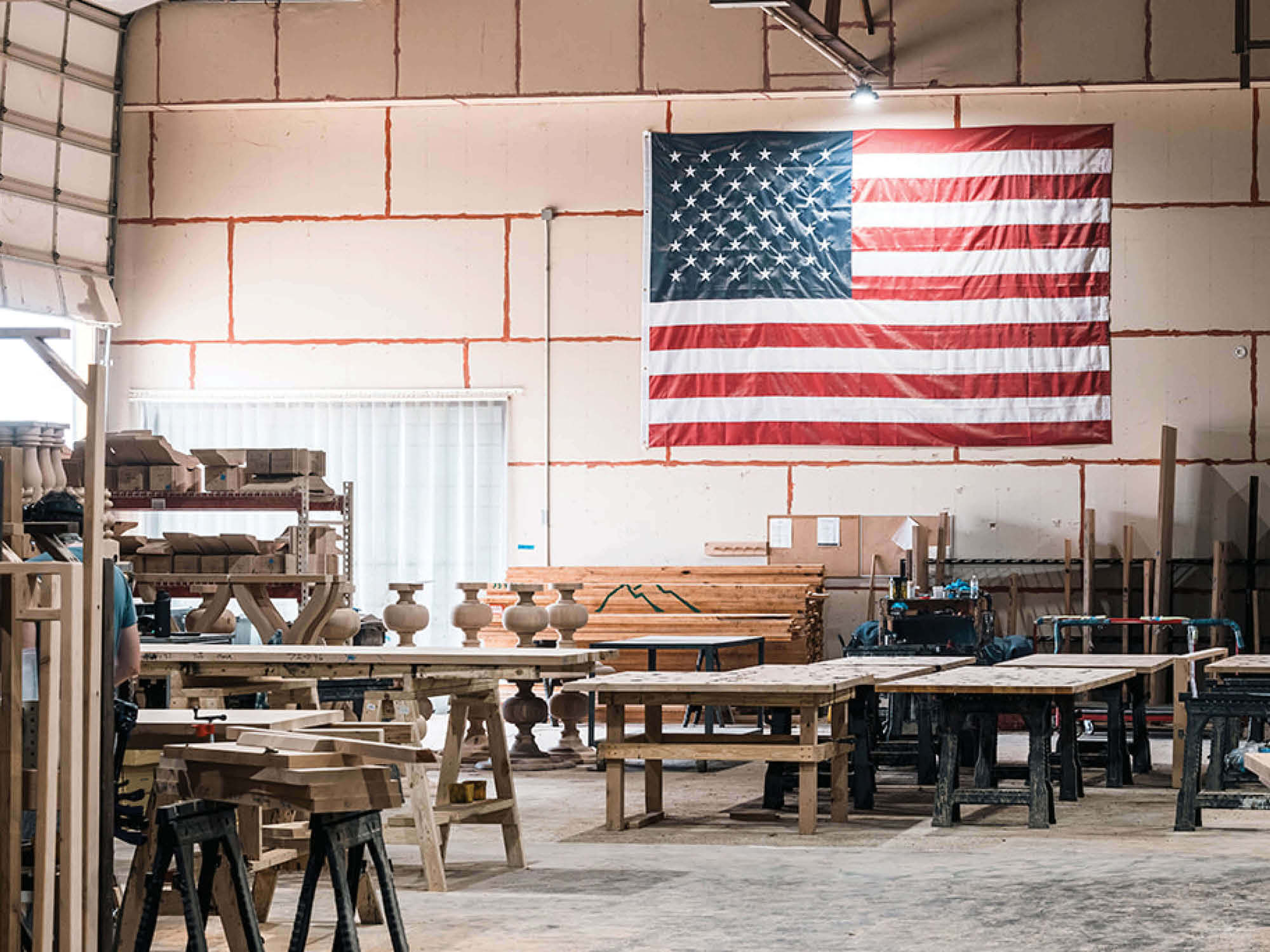 Interior of a woodworking shop with tables, tools, and wooden pieces in progress. A large American flag hangs on the wall, illuminated by overhead lights, adding a patriotic touch to the industrial workspace.
