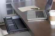 A tidy workspace with a wooden desk features a laptop, an open notebook, a smartphone, and a disposable coffee cup with a sleeve. Below the desk, the James+James Standard Desk Add-On—a keyboard tray with an empty compartment—is pulled out. Soft lighting enhances the clean and organized ambiance.