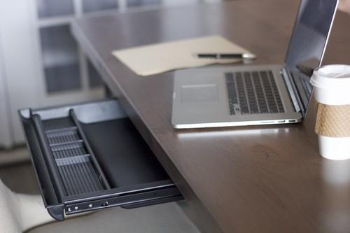 A tidy workspace with a wooden desk features a laptop, an open notebook, a smartphone, and a disposable coffee cup with a sleeve. Below the desk, the James+James Standard Desk Add-On—a keyboard tray with an empty compartment—is pulled out. Soft lighting enhances the clean and organized ambiance.