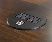 The James+James Standard Desk Add-On, a round black desk grommet featuring two standard power outlets and two USB ports, is embedded in a dark wooden surface.