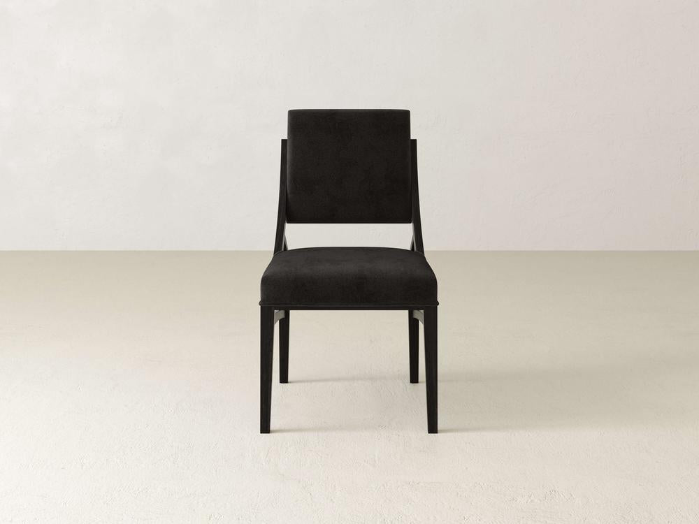 A black modern chair with a cushioned seat and backrest is centered against a plain, light-colored wall. The design is sleek and minimalist, with straight lines and a subtle, elegant appearance.