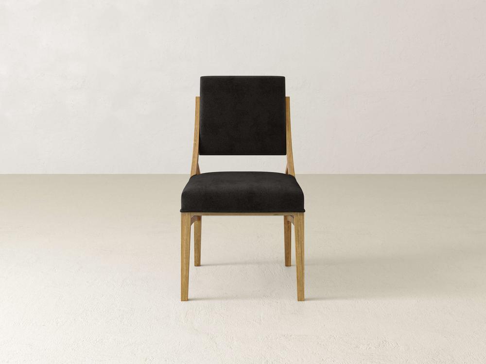 A minimalist wooden chair with a black cushioned seat and backrest is placed in the center of a plain, light-colored room with a smooth floor. The chair has a simple, modern design with clean lines.