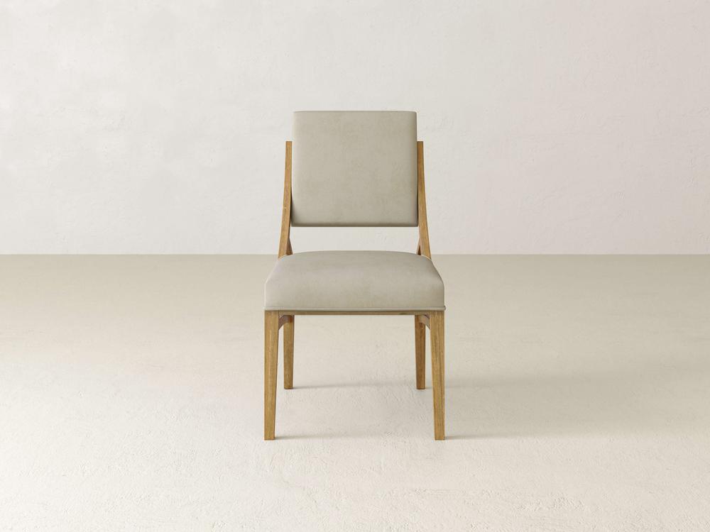 A modern wooden chair with a light beige padded seat and backrest is positioned on a plain, light-colored floor against a matching light-colored wall. The style is minimalist with clean lines.