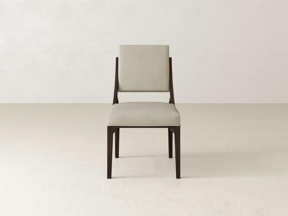 A modern, minimalist chair with a light beige cushioned seat and backrest. The chair features a dark wooden frame and is placed on a light-colored floor with a plain, neutral wall in the background.